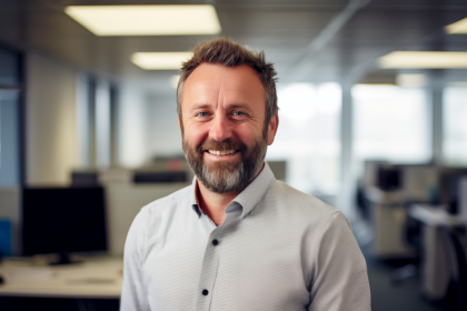Colin, a fleet manager in his 40s with light brown/greying hair and facial stubble. He is smiling at the camera while stood in an open plan office. There are banks of desks and computers behind him.