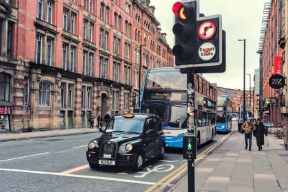 A cab and a bus, stopped at a traffic light on a Manchester street. Perspective view of large brick wall in background.