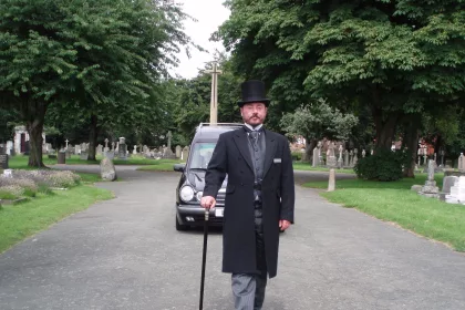 Funeral Director with a cane and top hat in front of a funeral car. Trees in the background.