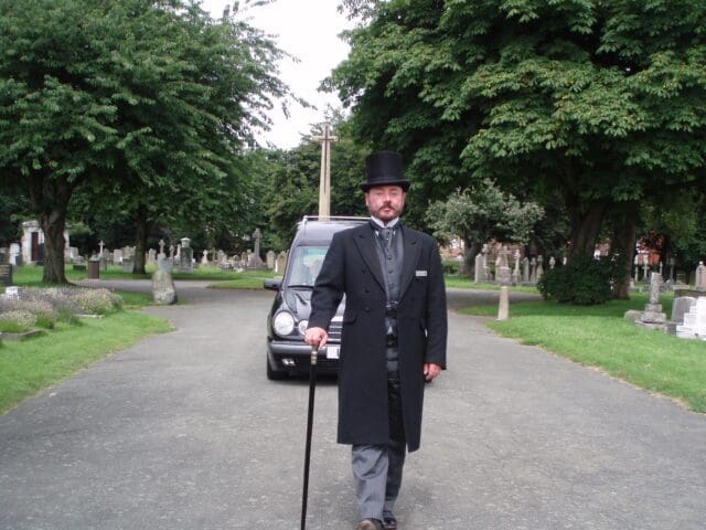 Funeral Director with a cane and top hat in front of a funeral car. Trees in the background.