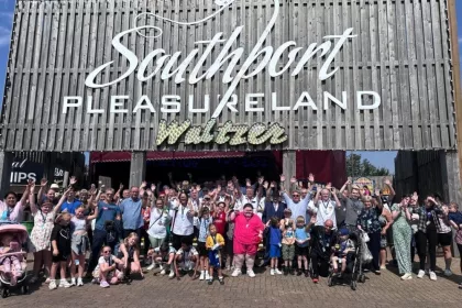 A group of over 40 taxi drivers stood with children outside Southport Pleasureland. They are all smiling and waving their hands in the air. This is Liverpool's 40th annual taxi outing, where taxi drivers volunteer to take vulnerable children on an all-expenses-paid day out of fun.
