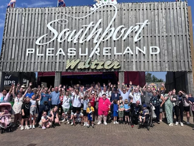 A group of over 40 taxi drivers stood with children outside Southport Pleasureland. They are all smiling and waving their hands in the air. This is Liverpool's 40th annual taxi outing, where taxi drivers volunteer to take vulnerable children on an all-expenses-paid day out of fun.