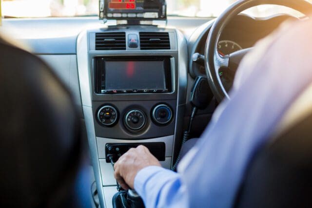 Taxi driver in vehicle, taken from behind. Radio and wheel are in view.
