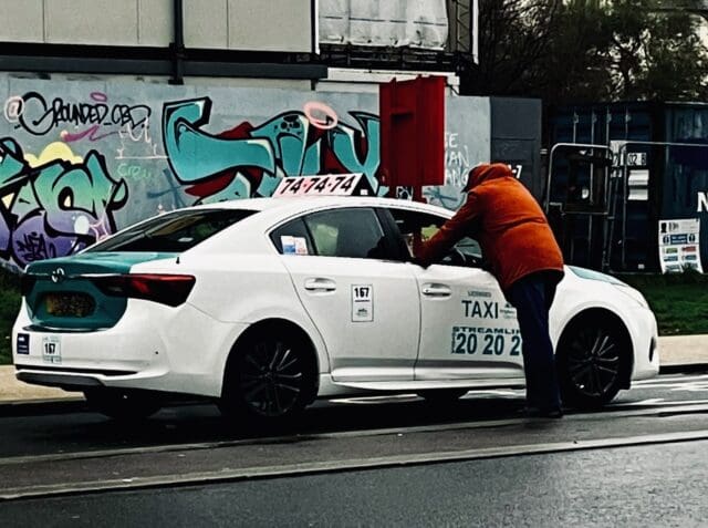man in orange jacket leaning into the window of a white private hire taxi