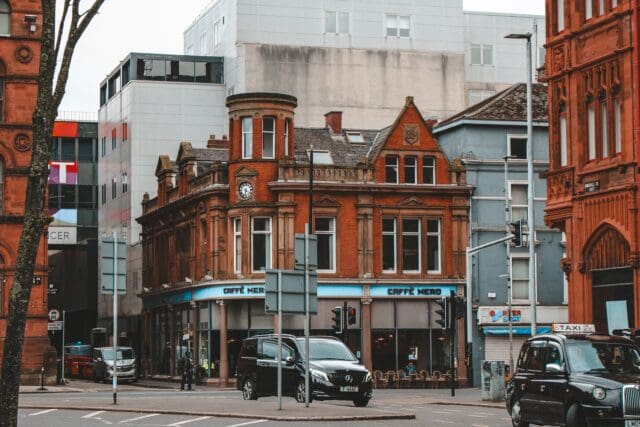 Street view of Belfast city featuring a historic red-brick building with ornate architectural details, housing a Caffè Nero on the ground floor. A black taxi and other vehicles are visible on the road, with modern buildings in the background. Traffic lights and street signs are present, reflecting an urban cityscape
