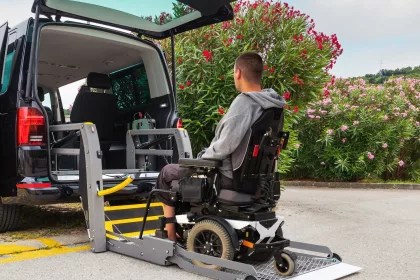 A man in a wheelchair is positioned onto an electric ramp attached to the back of a black taxi, waiting to be lifted into the cab.