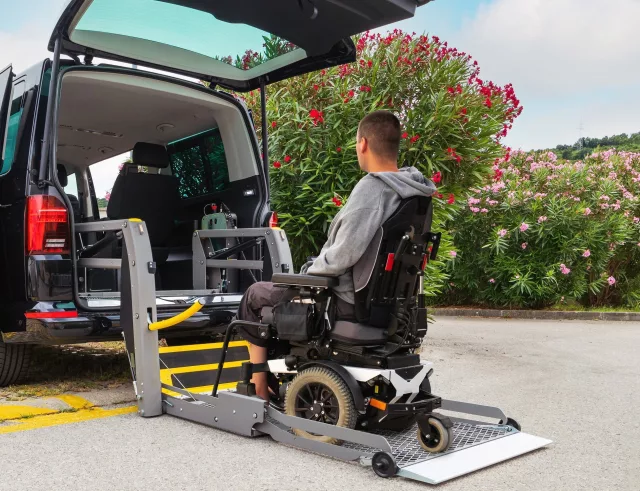 A man in a wheelchair is positioned onto an electric ramp attached to the back of a black taxi, waiting to be lifted into the cab.