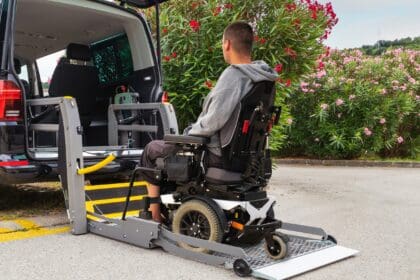 A man in a wheelchair is positioned onto an electric ramp attached to the back of a black taxi, waiting to be lifted into the cab.