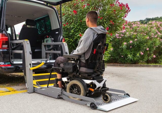 A man in a wheelchair is positioned onto an electric ramp attached to the back of a black taxi, waiting to be lifted into the cab.
