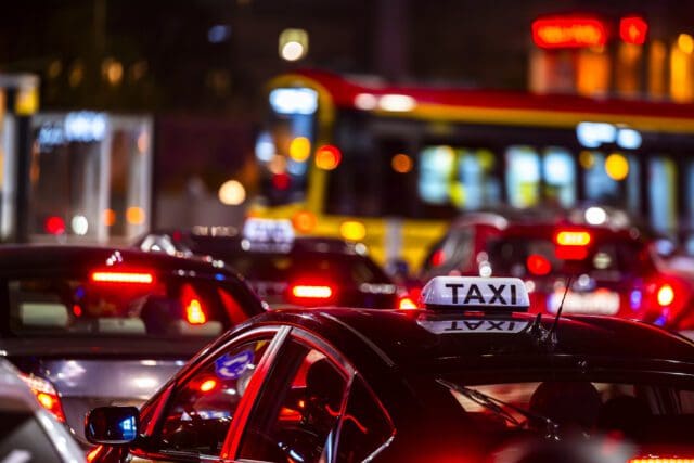 A night-time urban scene featuring a congested traffic jam with various vehicles. A black car in the foreground has a lit "TAXI" sign on its roof, while bright red brake lights and streetlights illuminate the surroundings.
