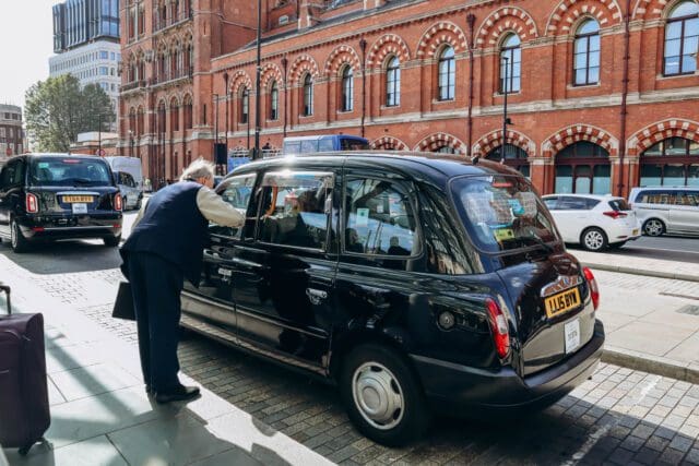 A black London taxi is parked near a red-brick building with arched windows in London. A man wearing a dark vest and trousers leans into the open passenger window, engaging in conversation with the driver. The background features other black taxis, pedestrians, and modern buildings.