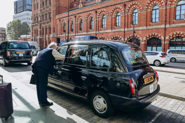 A black London taxi is parked near a red-brick building with arched windows in London. A man wearing a dark vest and trousers leans into the open passenger window, engaging in conversation with the driver. The background features other black taxis, pedestrians, and modern buildings.