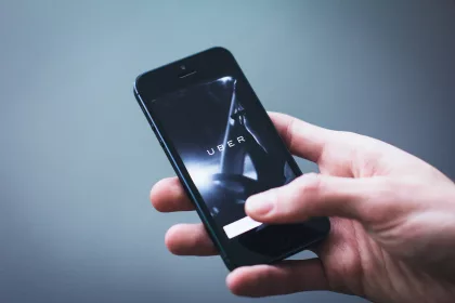 A person holding a smartphone displaying the Uber app on the screen, with a dark background and a hand interacting with the device.