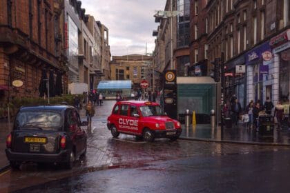 A city street scene in Glasgow, Scotland, featuring two traditional London-style taxis—one black and one red—on a wet, reflective cobblestone road. Pedestrians are walking along the street. The background consists of a mix of modern and historic sandstone buildings. The sky is overcast, and the street appears busy with people carrying shopping bags.