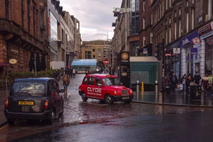 A city street scene in Glasgow, Scotland, featuring two traditional London-style taxis—one black and one red—on a wet, reflective cobblestone road. Pedestrians are walking along the street. The background consists of a mix of modern and historic sandstone buildings. The sky is overcast, and the street appears busy with people carrying shopping bags.