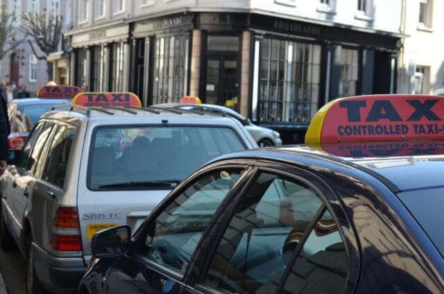 A fleet of taxis parked along a street in an urban setting during daytime. The taxis have illuminated roof signs that read 'TAXI CONTROLLED TAXI-CAB' in bold red and black lettering with yellow accents.