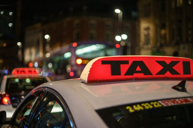 A close-up of a red illuminated taxi sign on the roof of a white car at night. There is a dark urban background, with features blurred city lights, buildings, and another taxi with a similar sign. A bus with bright headlights is visible in the background.