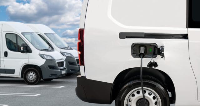A white electric van is charging at a charging station, with a cable plugged into its charging port. A green battery indicator is visible on the charging panel. In the background, two other similar white delivery vans are parked in a row under a partly cloudy sky.