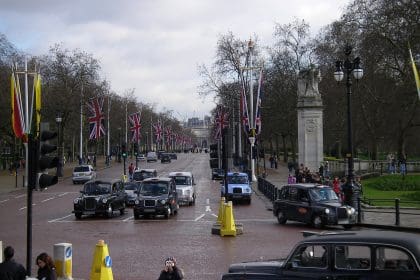 A busy street scene in London, featuring multiple black cab taxis and other vehicles on a wide road lined with Union Jack flags. Pedestrians are walking along the sidewalks, some taking photos, while others are gathered near a monument.