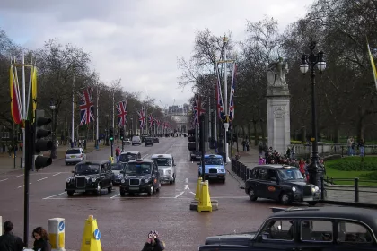 A busy street scene in London, featuring multiple black cab taxis and other vehicles on a wide road lined with Union Jack flags. Pedestrians are walking along the sidewalks, some taking photos, while others are gathered near a monument.