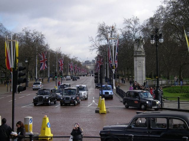A busy street scene in London, featuring multiple black cab taxis and other vehicles on a wide road lined with Union Jack flags. Pedestrians are walking along the sidewalks, some taking photos, while others are gathered near a monument.