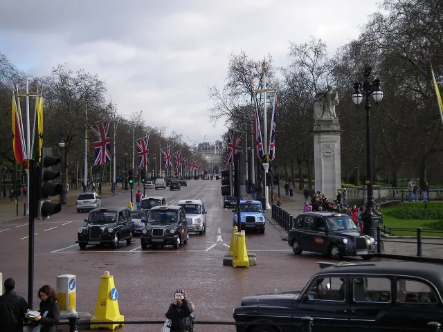 A busy street scene in London, featuring multiple black cab taxis and other vehicles on a wide road lined with Union Jack flags. Pedestrians are walking along the sidewalks, some taking photos, while others are gathered near a monument.
