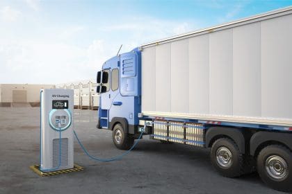 3D rendering of an electric heavy goods lorry (HGV) in a logistics yard is connected to an EV charging station. The blue and white truck is parked beside a modern charging unit with a digital display and glowing circular light, indicating active charging. The background features a row of loading docks and a clear blue sky.