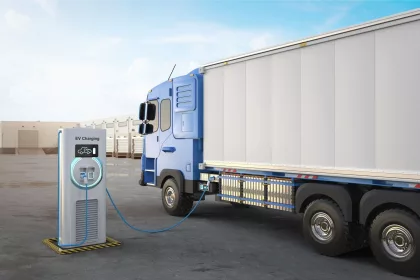 3D rendering of an electric heavy goods lorry (HGV) in a logistics yard is connected to an EV charging station. The blue and white truck is parked beside a modern charging unit with a digital display and glowing circular light, indicating active charging. The background features a row of loading docks and a clear blue sky.