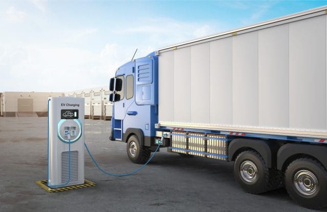 3D rendering of an electric heavy goods lorry (HGV) in a logistics yard is connected to an EV charging station. The blue and white truck is parked beside a modern charging unit with a digital display and glowing circular light, indicating active charging. The background features a row of loading docks and a clear blue sky.