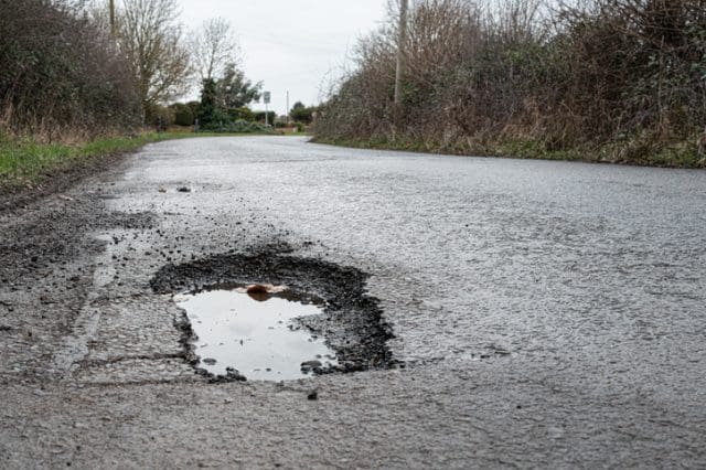 A large pothole filled with water on a rural British road, surrounded by broken asphalt and loose gravel. The road is damp and bordered by overgrown hedges and leafless trees under an overcast sky. The scene is quiet and slightly dreary, typical of a countryside lane in late autumn or winter.