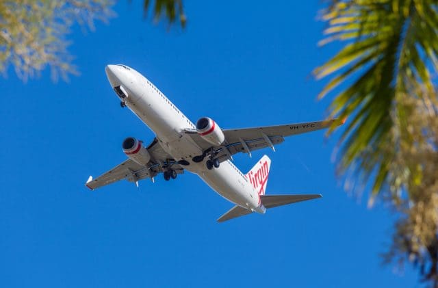 Virgin Atlantic airplane flying overhead on a clear blue sky day, framed by tropical palm trees.