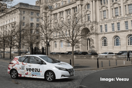 A Veezu taxi parked in front of the Liver building in Liverpool. Leafless trees line the sidewalk, and a few pedestrians are visible in the background. The sky is overcast.