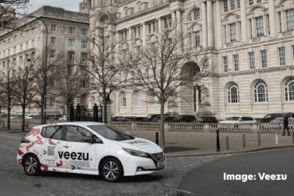 A Veezu taxi parked in front of the Liver building in Liverpool. Leafless trees line the sidewalk, and a few pedestrians are visible in the background. The sky is overcast.