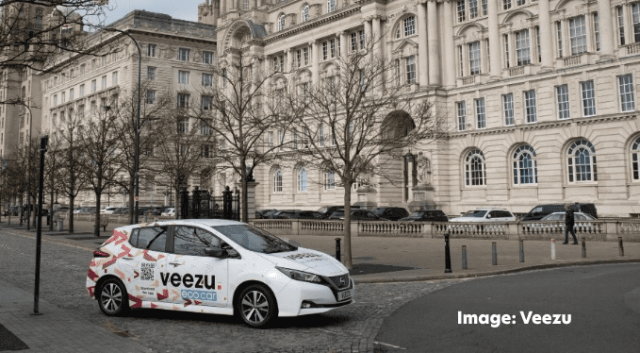 A Veezu taxi parked in front of the Liver building in Liverpool. Leafless trees line the sidewalk, and a few pedestrians are visible in the background. The sky is overcast.