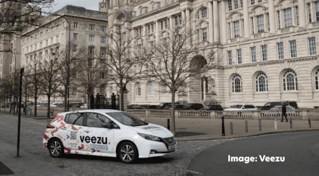 A Veezu taxi parked in front of the Liver building in Liverpool. Leafless trees line the sidewalk, and a few pedestrians are visible in the background. The sky is overcast.