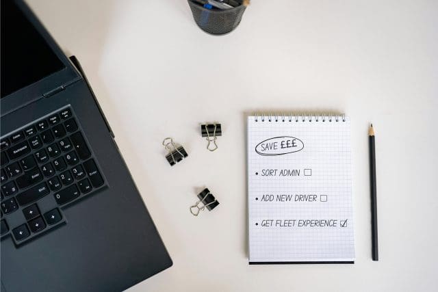 A white desk which has a laptop keyboard to its left, with three paper clips in the middle and a notebook and pencil on its right. The note book has a to-do list that reads: Save money, sort admin, add new driver, get fleet experience.