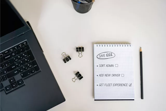 A white desk which has a laptop keyboard to its left, with three paper clips in the middle and a notebook and pencil on its right. The note book has a to-do list that reads: Save money, sort admin, add new driver, get fleet experience.