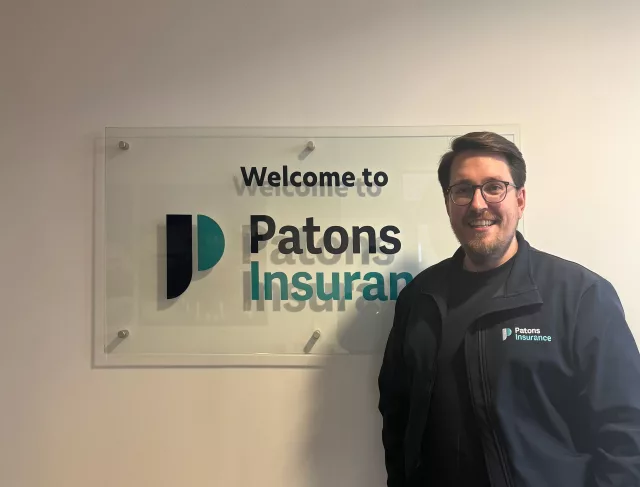 Patons Insurance marketing manager, Ben, pictured smiling next to Patons Insurance logo signage