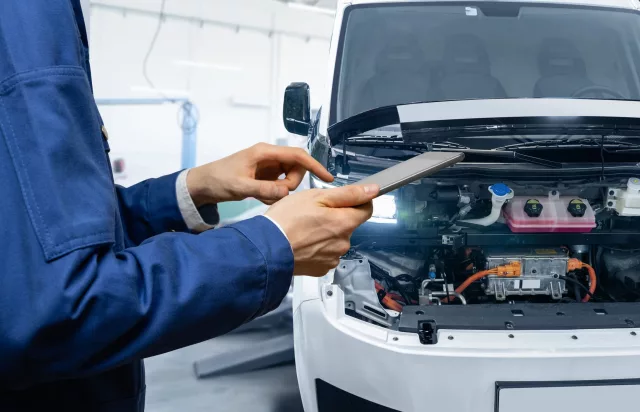 Serviceman with digital tablet next to electric van with open hood. EV maintenance.