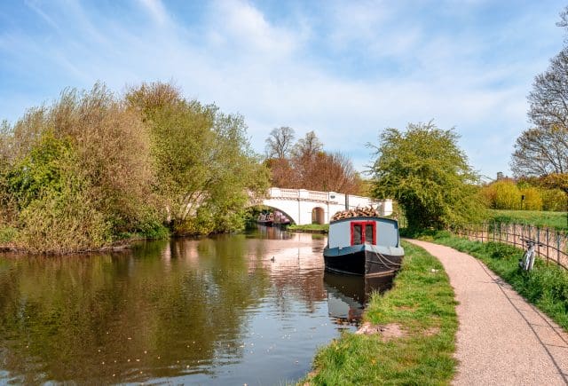 View of the river Gade (part of Grand Union Canal) and The Grove Bridge (Grove Ornamental Bridge No 164) in Cassiobury Park, Watford, England. Moored narrow boat, reflections on water.