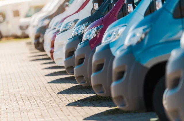 Fleet of vans in different colors sit neatly in a parking lot under a clear sky, highlighting their distinctive designs and shapes.