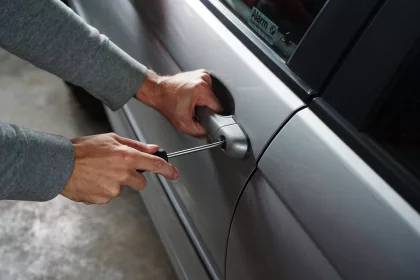 Close-up of a person attempting to break into a silver car using a screwdriver on the door handle lock.