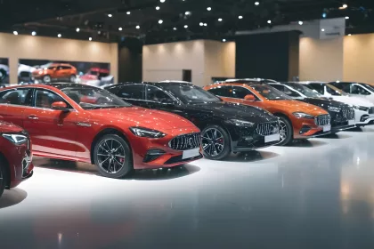 A brightly lit modern car dealership showroom displaying a lineup of cars in red, black, orange, and white. The vehicles are parked side by side on a glossy showroom floor under ceiling spotlights.