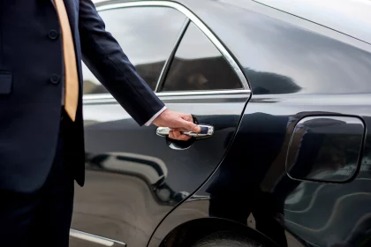 Chauffeur in a dark suit and light-colored tie is opening the rear door of a black luxury car, with their hand on the door handle and the vehicle’s side visible in close-up.