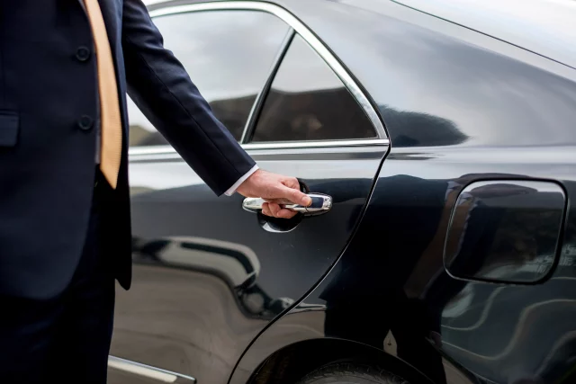 Chauffeur in a dark suit and light-colored tie is opening the rear door of a black luxury car, with their hand on the door handle and the vehicle’s side visible in close-up.