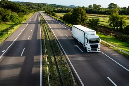A white articulated lorry with a curtain-sided trailer travels along a dual carriageway, photographed from an elevated angle. The road stretches into the distance through a green, rural landscape with trees, fields, and hedgerows on either side under a clear sky.