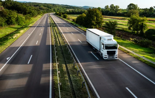 A white articulated lorry with a curtain-sided trailer travels along a dual carriageway, photographed from an elevated angle. The road stretches into the distance through a green, rural landscape with trees, fields, and hedgerows on either side under a clear sky.