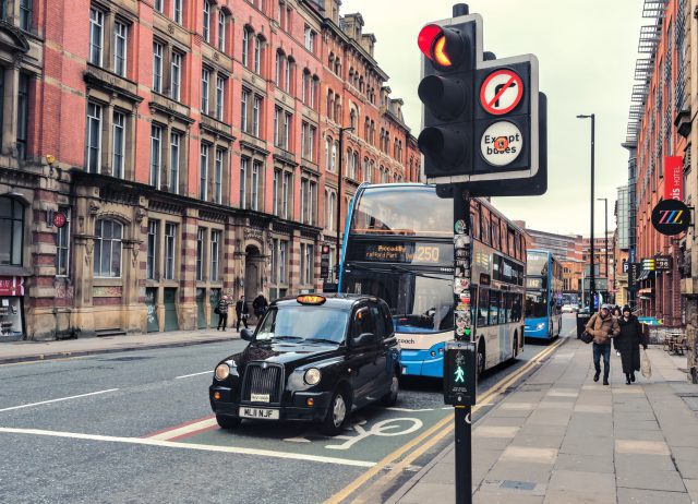 A cab and a bus, stopped at a traffic light on a Manchester street. Perspective view of large brick wall in background.